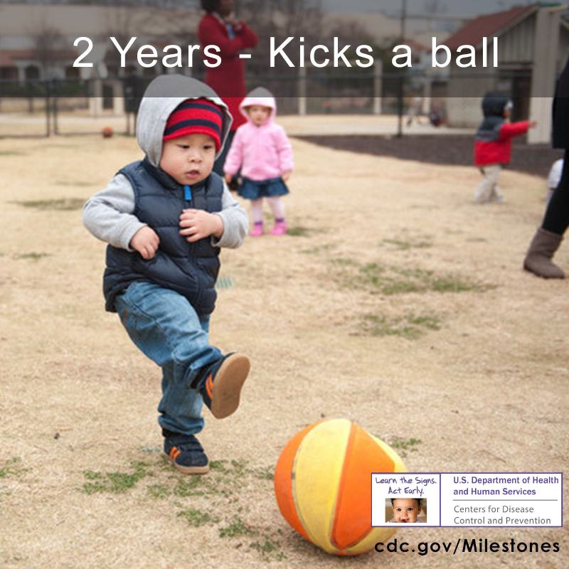 The boy in this photo is kicking a yellow and orange ball, a 2 year milestone.