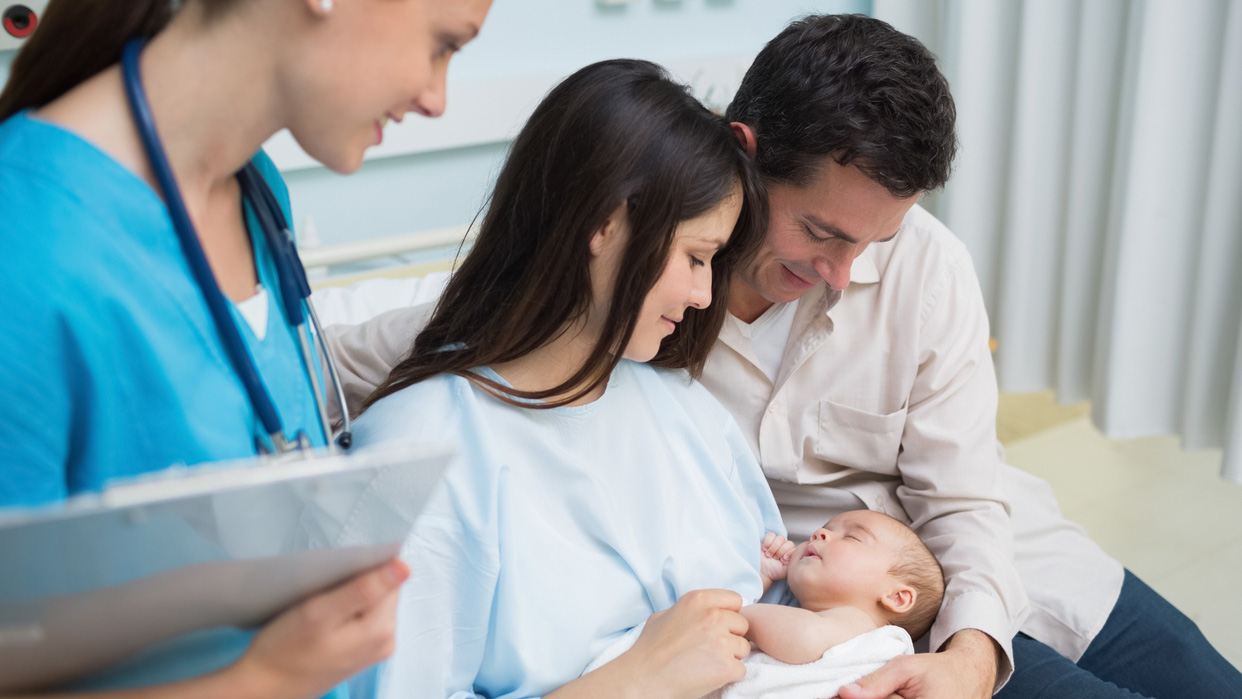 mPINC FAQ Doctor looks at a mom and dad holding a baby in diaper in a hospital room.