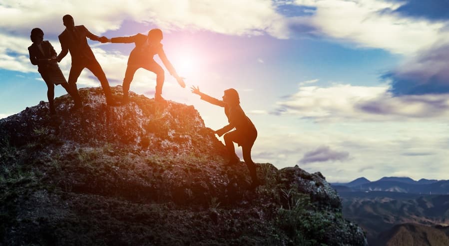 Photo of three people at the top of a mountain helping a fourth person climb up