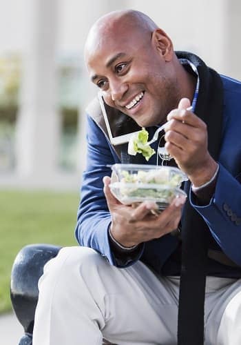 Photo of a man eating a salad while sitting on a park bench talking on his cell phone