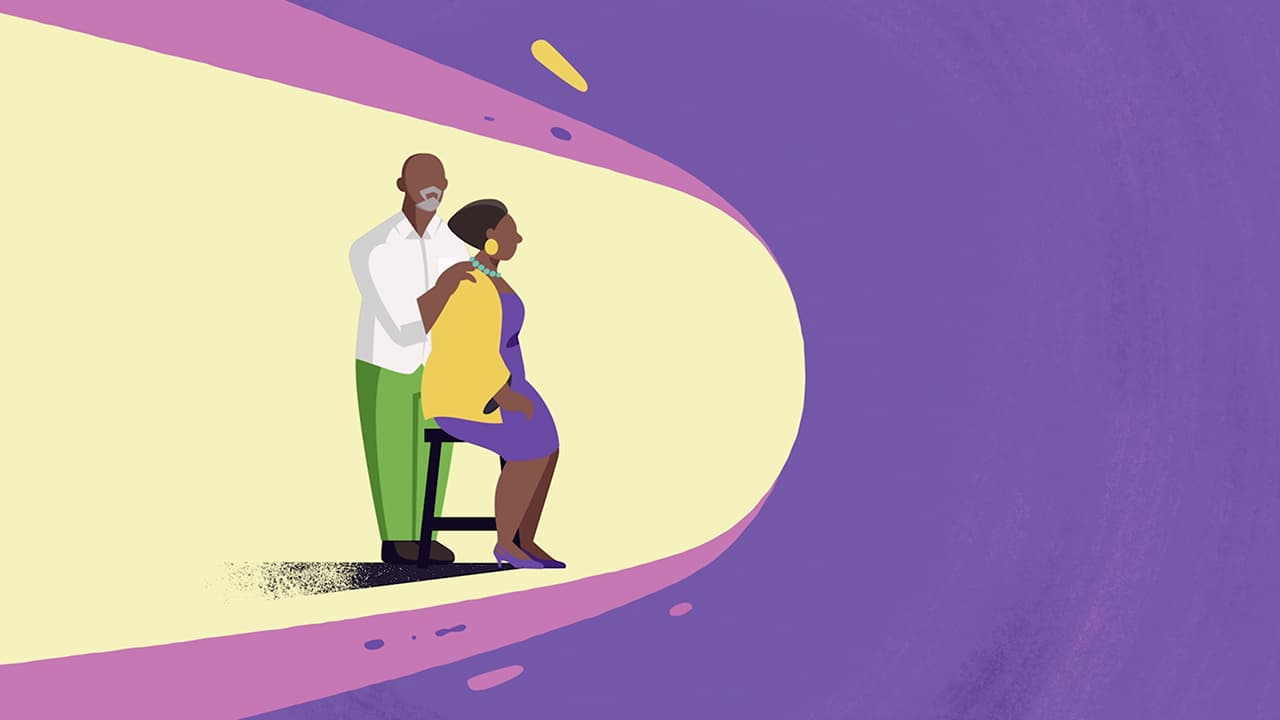 Linda sits on a stool and covers her face with her hands while her husband stands behind her and holds her shoulders.