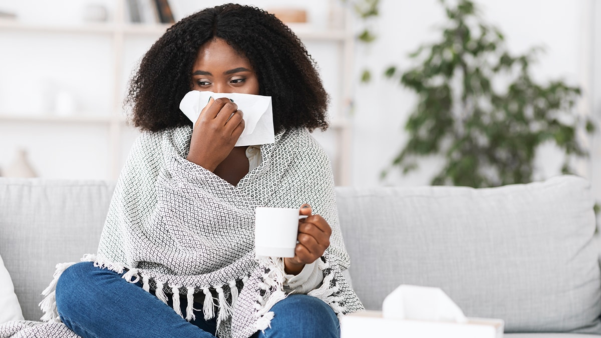 Woman treating her cold Sick woman on a couch draped in a blanket drinking tea and treating her cold symptoms.