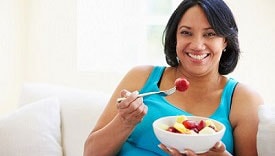 Overweight Woman Sitting On Sofa Eating Bowl Of Fresh Fruit Woman Sitting On Sofa Eating Bowl Of Fresh Fruit