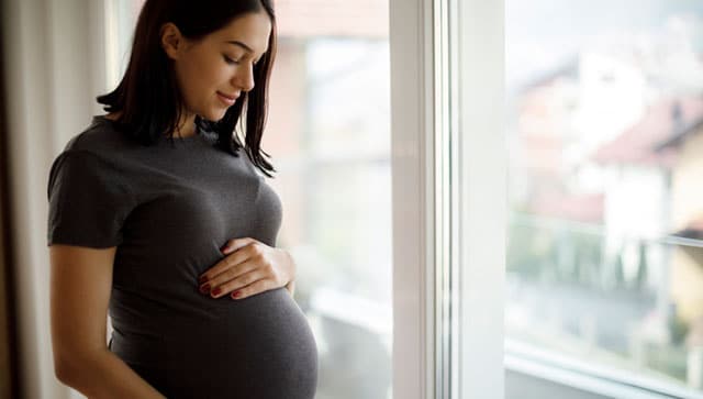 Portrait of young happy pregnant woman standing by the window Pregnant woman looks down at her belly.