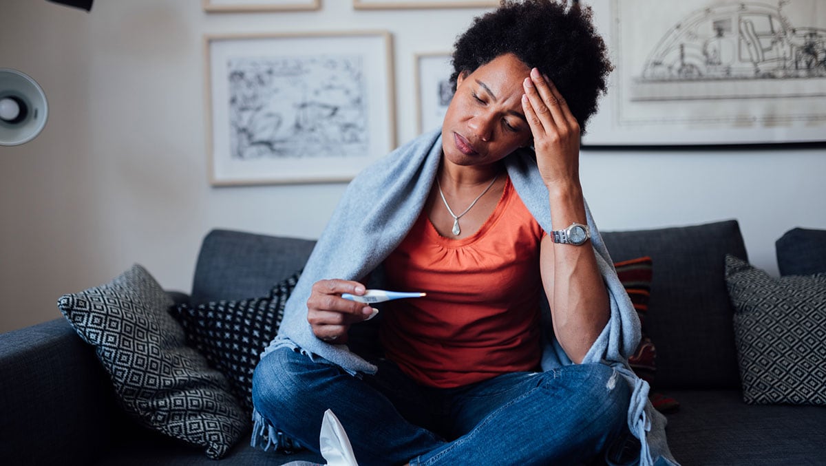 Woman sitting on couch with a blanket, looking at a digital thermometer.