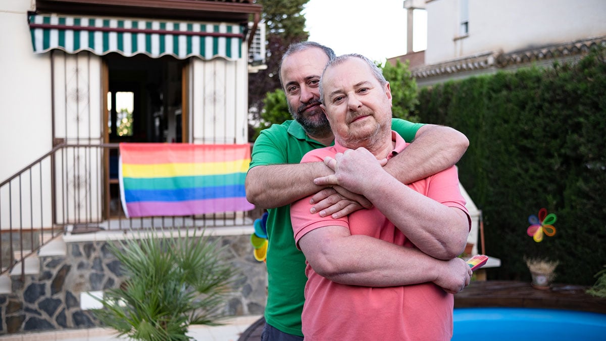 Middle-aged couple embracing in the garden of their home with an LGBT flag in the background. middle aged LGBTQ couple