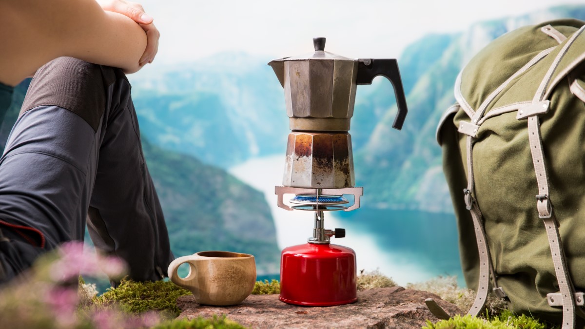 Sitting hiker looking out over mountains and water. Next to the hiker are a mug, small kettle boiling over a gas burner, and a backpack.