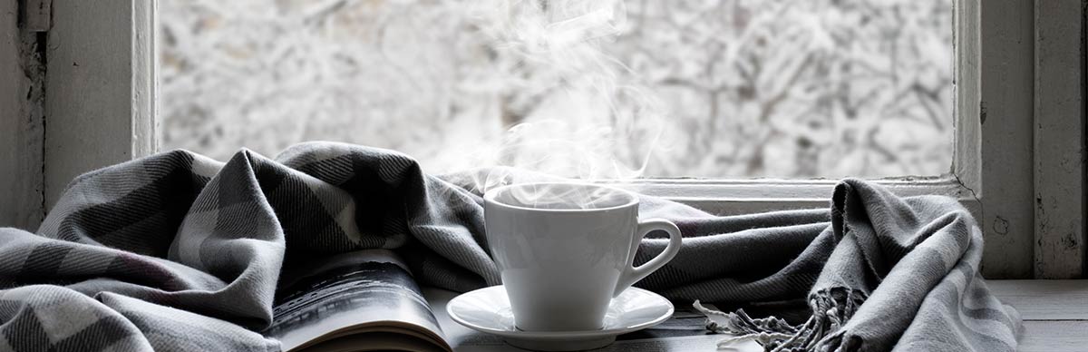 Un libro, una bufanda gris y blanca y una taza blanca llena de una bebida caliente y humeante se colocan en una mesa frente a una ventana que muestra un paisaje nevado afuera. Un libro, una bufanda gris y blanca y una taza blanca llena de una bebida caliente y humeante se colocan en una mesa frente a una ventana que muestra un paisaje nevado afuera.