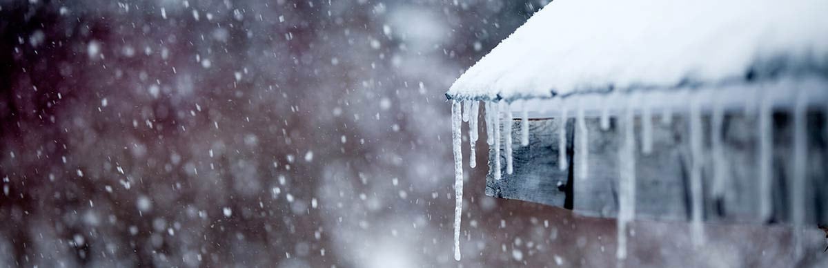 winter-weather-prepare-home Techo cubierto de nieve con carámbanos a lo largo del borde y nieve cayendo.
