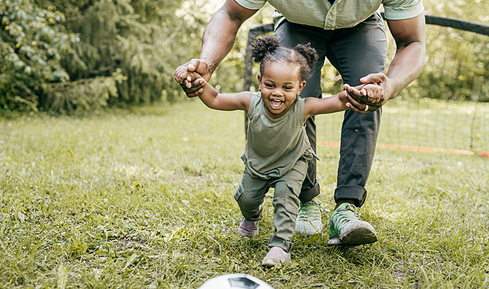 Caregiver helping a toddler run towards a soccer ball. Caregiver helping a toddler run towards a soccer ball.