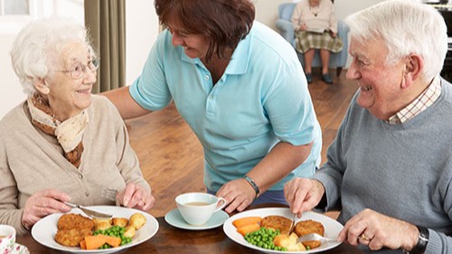 Staff member checking on elderly couple in assisted living dining room.