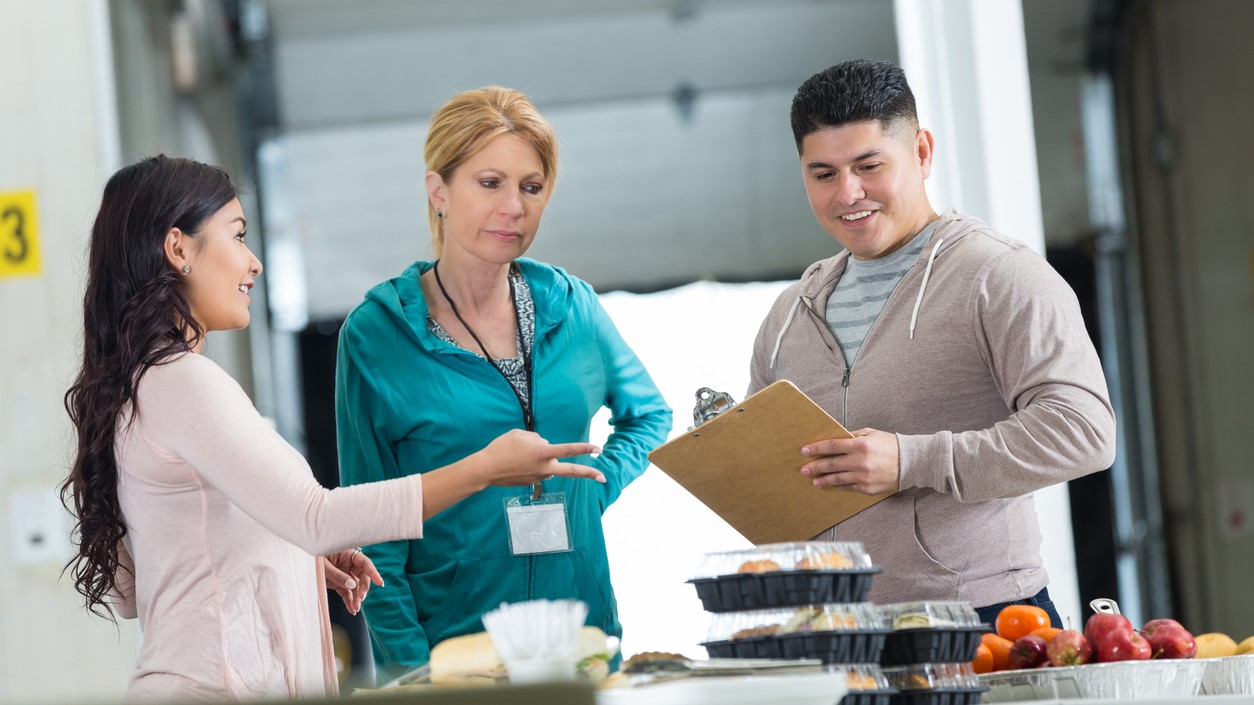 Three adults talking by a food service counter. One has a clipboard, and one is wearing a badge.