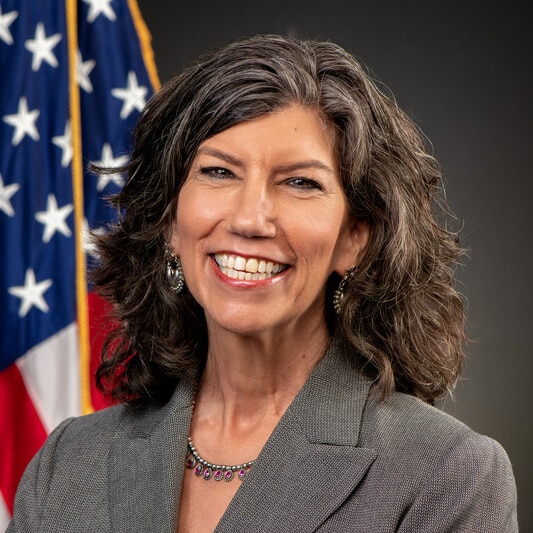 Dr. Kayla Laserson, GHC Director Woman with dark gray hair against a gray background with the U.S. flag.