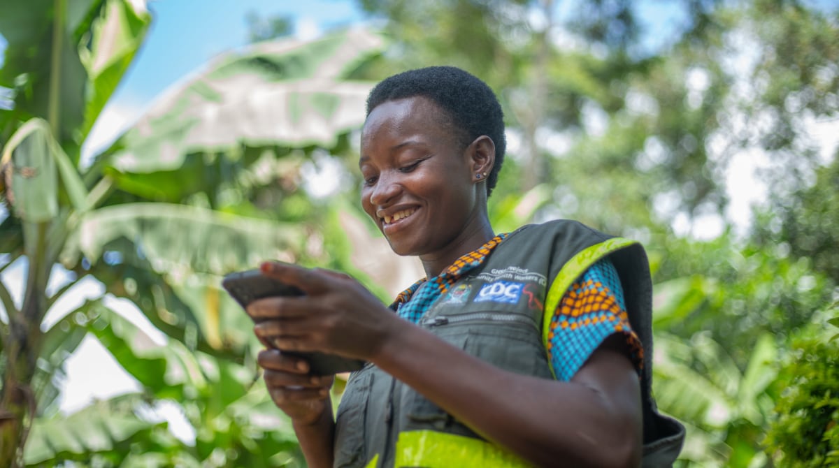 Tanzania Vedastina Shumbusho Woman wearing a CDC vest smiles as she holds electronic device.