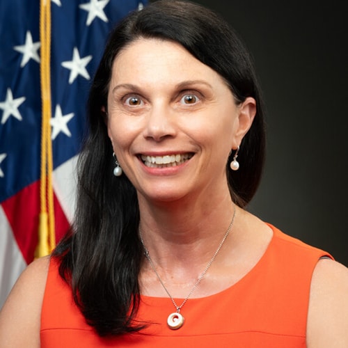 A professional headshot of a woman with long dark hair, wearing a bright orange sleeveless dress and pearl earrings. She is smiling widely, and behind her, there is an American flag.