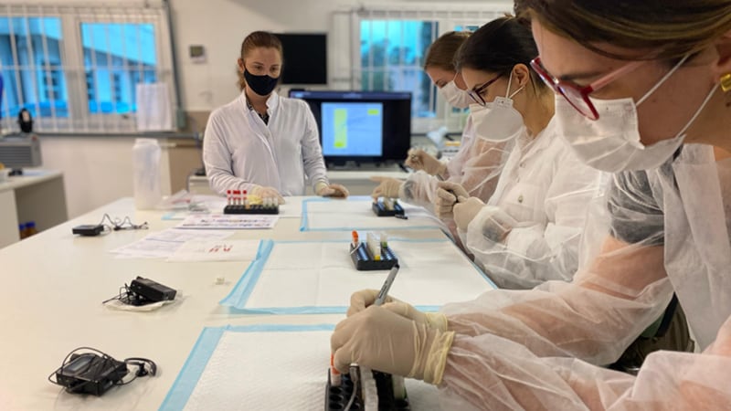 Recency Training 2022 Lab technicians in masks and gloves analyze samples at a lab bench.
