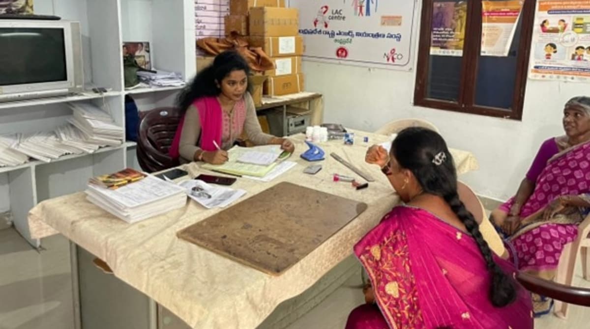Narisaksham HIV Clinic in India A woman in a health clinic sits at a table while she fills paperwork. She is engaging with two other women seating across the table. The room contanains various files and suplies around.
