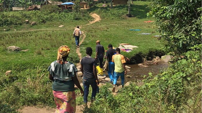 DRC_Ebola_FieldPhoto A group of people in the grassy plains, crossing a small stream with stone pathing, along a path.