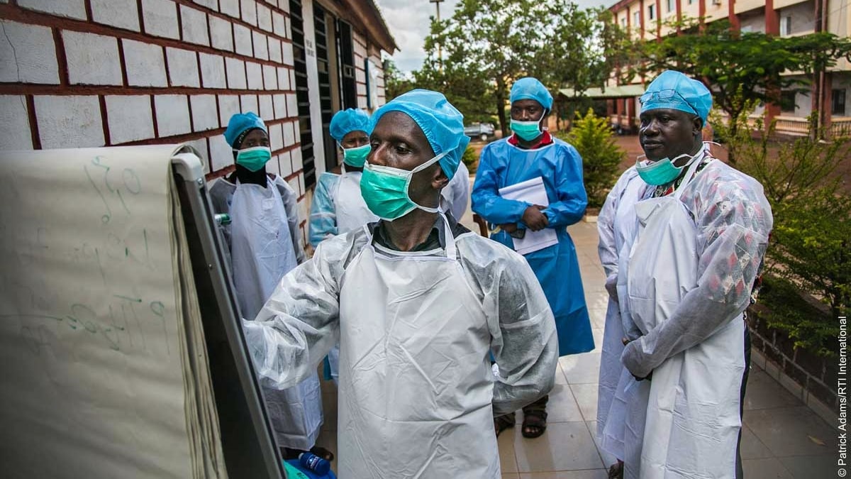 RTI_Guinea_Epidemiology_Training_Photo A health worker writes on a flip board. Four other health workers stand behind him. All are wearing full personal protective equipment.