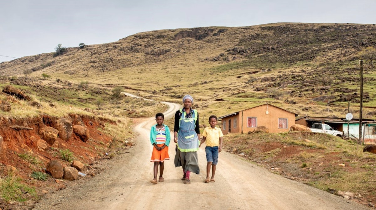 South Africa Main Image A woman with a young boy and girl beside her on road. Behind them is a house.
