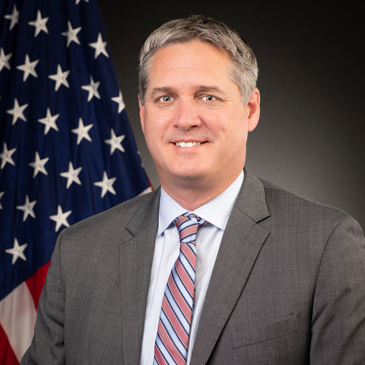 A man with gray hair smiles while wearing a light blue shirt, gray suit jacket, and a red and blue striped tie.