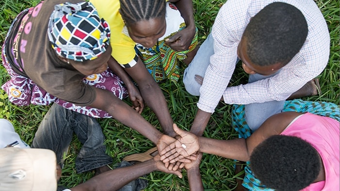 mozambique-16x9 A Community group boosts access to HIV treatment, sitting in a circle, hand stacked, middle hand on top holding a pill