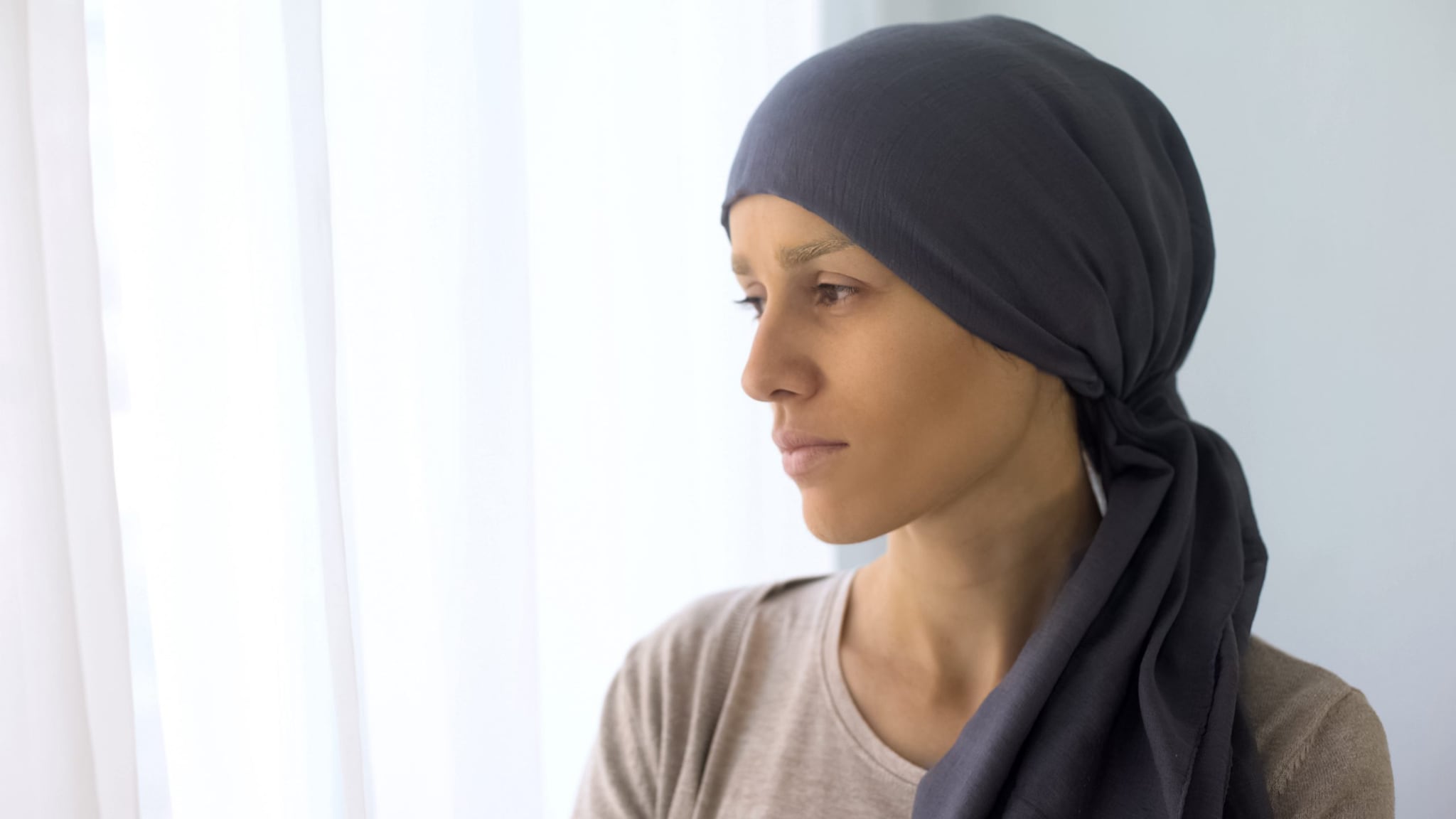 Upset woman in headscarf looking in window, rehabilitation centre, fatal disease Photo of a woman with cancer, wearing a head covering, looking off into the distance.