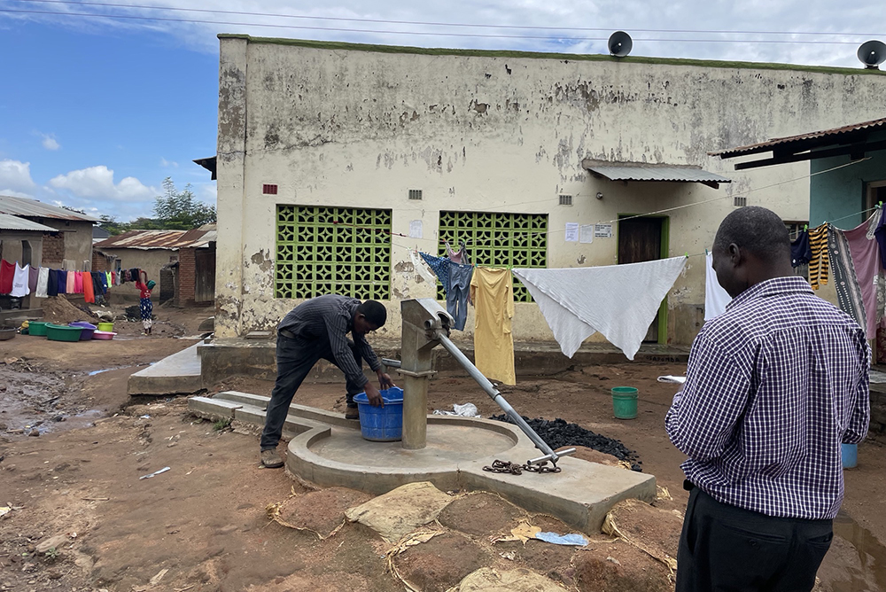 man filling tub with water at outdoor pump while another man looks on