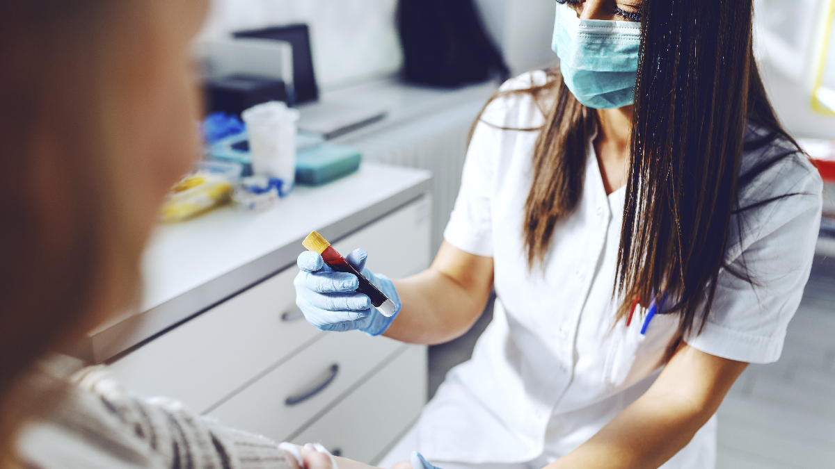 OccupationalExposure Woman in a medical setting, wearing a white medical uniform and a mask, holding a vial of blood.