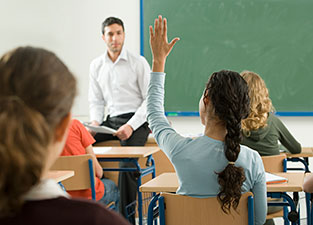 Student holding up her hand; teacher sits in front of classroom