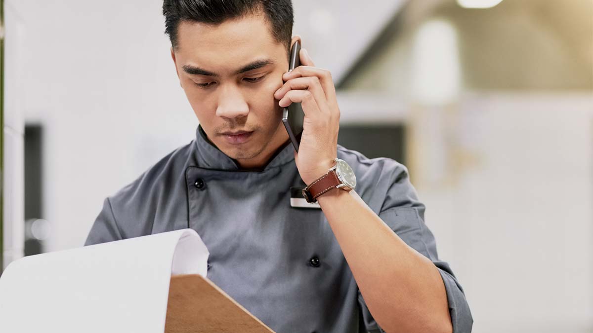 This figure is an image of a restaurant worker taking a phone call and looking at a clipboard.
