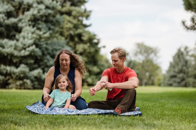 A couple with a young girl sitting on a blanket on the grass. The man is applying insect repellant on his arm.