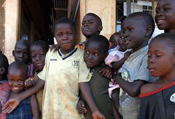 This photograph shows a group of young African children standing side by side. A few are looking at the camera and smiling while others are looking to the side. 