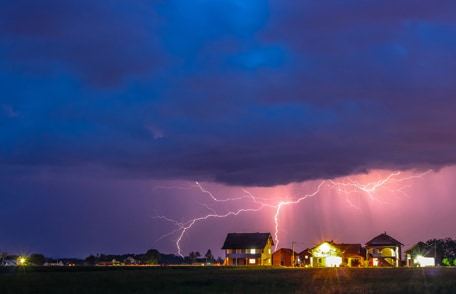 A large storm with lots of lightning approaches a neighborhood.