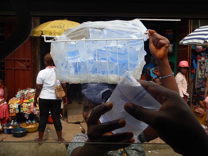 A woman walks through a marketplace carrying a basket on her head full of plastic pouches that contain water 