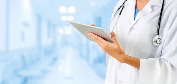 Doctor writing A doctor in a white coat with a stethoscope around their neck uses a tablet to review a report in a hospital hallway.