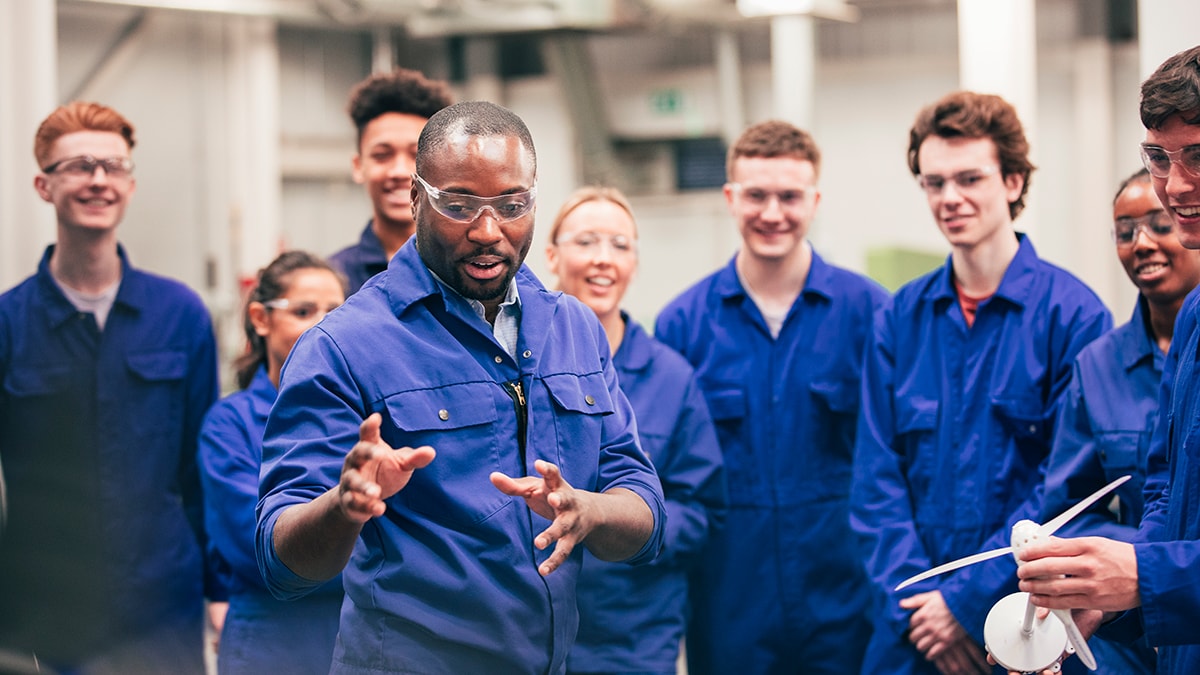 — title missing — Group of people wearing coveralls watching someone give a demonstration.
