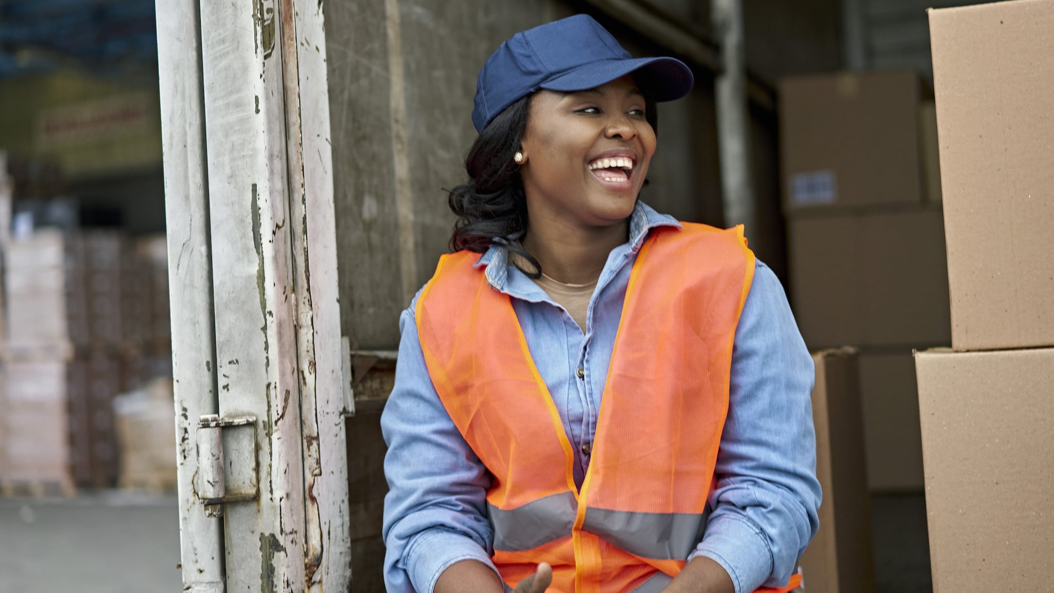 — title missing — woman wearing blue baseball hat and orange safety vest sitting on the back of a truck with boxes