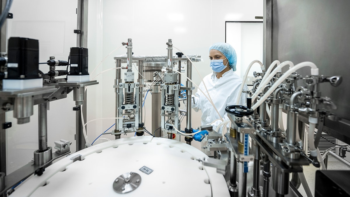 — title missing — Small brown bottles seen in a laboratory machine during manufacturing in a pharmaceutical factory while a female employee in a protective clothing is controlling the process.
