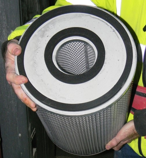 A mine worker holding the filter before it is to be installed in the filter housing, with the angle of the photo showing the double gasket seal near the filter?s smaller inner diameter and filter?s larger outer diameters. The filter is approximately eight inches in diameter and twelve inches long.