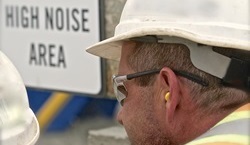 hlp.jpg A construction worker wearing hearing protection in front of a sign which reads "High Noise Area"