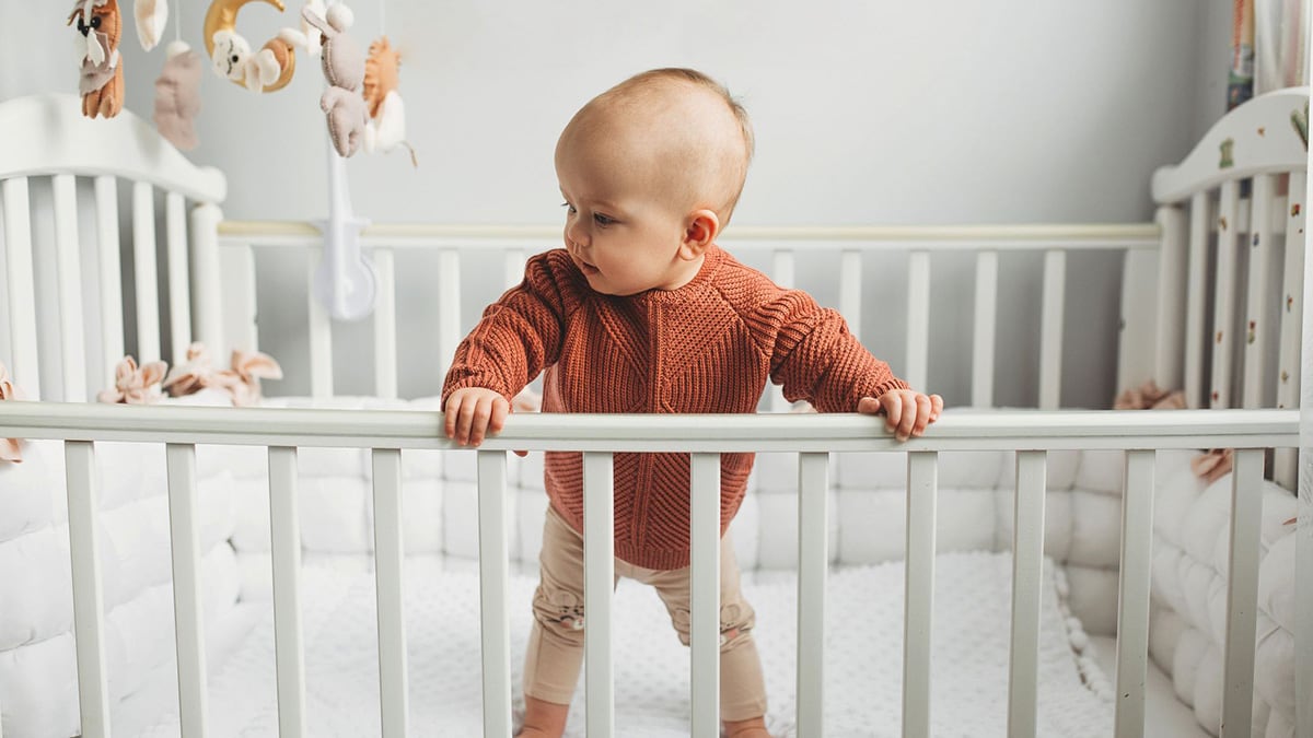 Toddler in a crib Beautiful little girl in a crib in a cozy light room in a sweater of fashionable shades. learns to stand at the support.