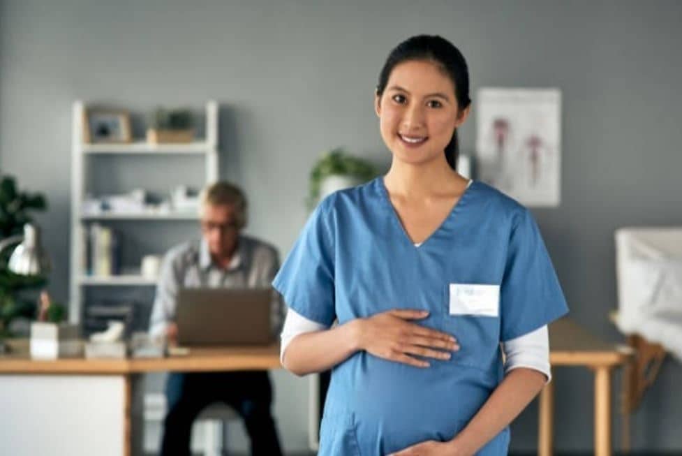 parvovirus-pregnancy-workplace Pregnant woman wearing scrubs and holding her belly with a doctor in a white coat sits in the background at the computer