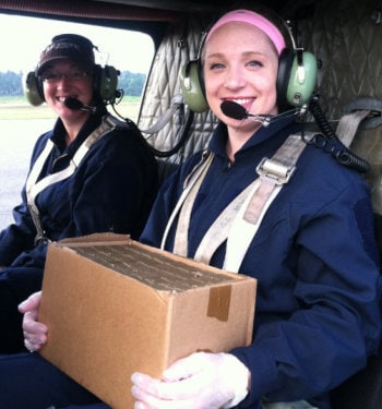 Katherine Mullican Two young women in a helicopter, one of them wearing gloves and carrying a sealed box of supplies.