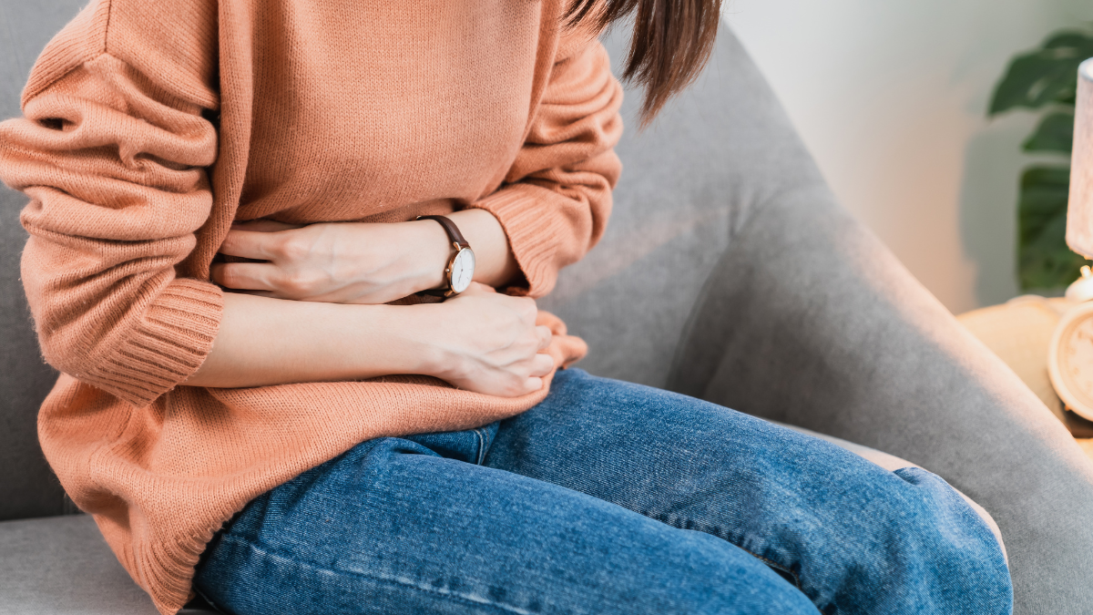 Cramps A person sitting down hunched over on a gray chair, holding their stomach with both hands.