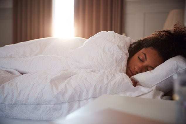 Woman sleeping in bed as day breaks through closed curtains.