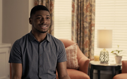 Black male sitting in room with window and chair behind him talking to camera