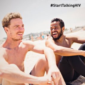 summer Image displays two men smiling/laughing while sitting on a beach.