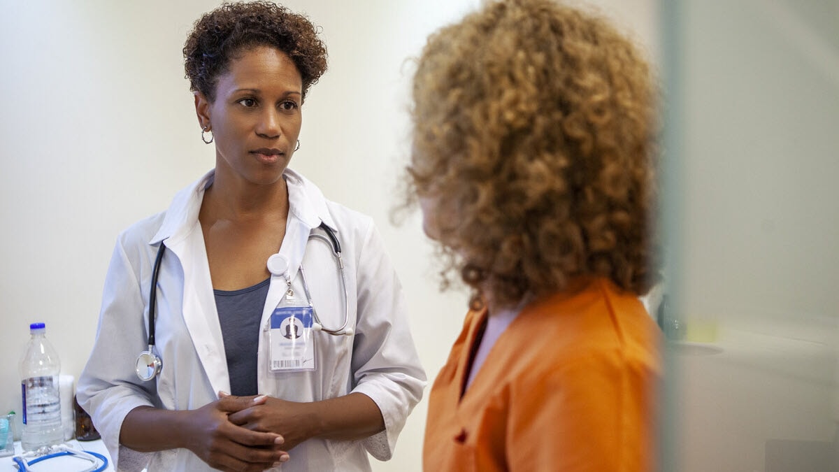 Mid shot of African-American female doctor talking to incarcerated woman wearing orange jumpsuit in correctional healthcare facility