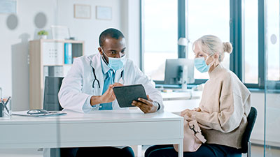African American Doctor in Protective Mask is Reading Medical History of Senior Female Patient During Consultation in a Health Clinic. Physician Using Tablet Computer in Hospital Office. Health care worker speaking to patient
